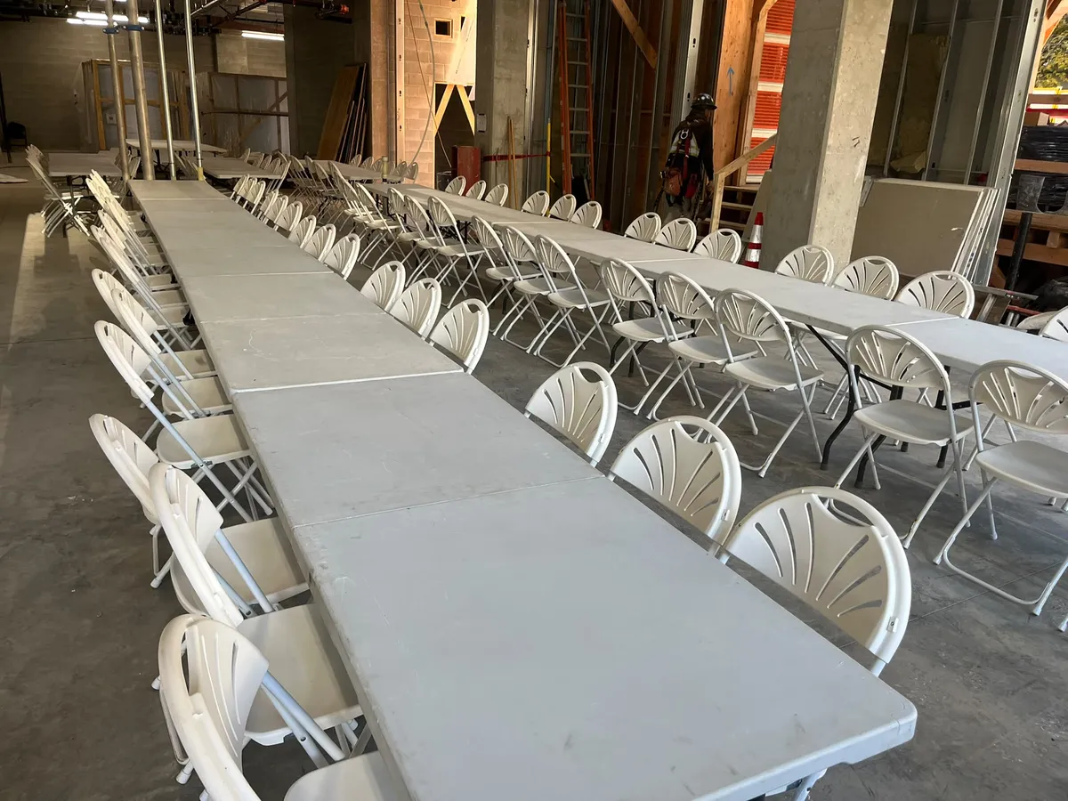 Long rows of banquet tables and white fanback chairs set for a crew lunch inside a building under construction
